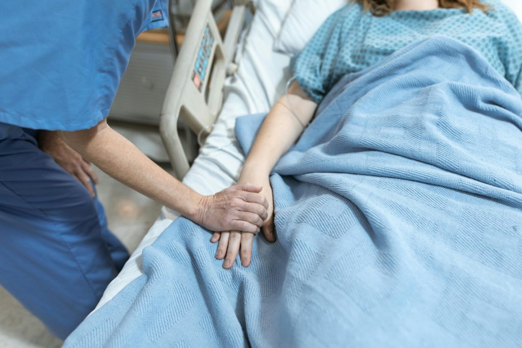 Ein Krankenbett, in dem ein Patient liegt. Eine Pflegekraft hält seine Hand